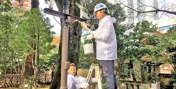 写真：白の作業着にヘルメット被った作業員がハシゴに登り、柱に塗料を塗っている様子。