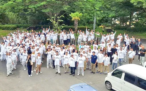 写真：白の作業着にヘルメット被った作業員たちが広場に集まり手を挙げている様子。