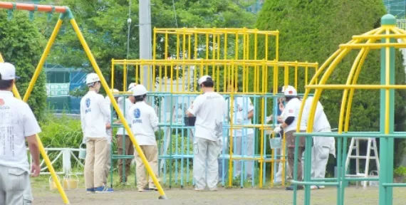 写真：白の作業着にヘルメット被った作業員が公園の遊具に複数人で塗料を塗っている様子。